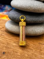 Gold cylindrical pendant with knurled ends and two engraved tubes inside, suspended from a circular bail atop stacked stones.