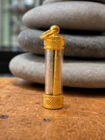 Gold cylindrical pendant with engraved tubes inside a clear glass capsule and a hanging ring on top, resting on a wooden surface.