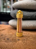 Gold-toned decorative glass capsule with textured gold caps, containing two engraved metal rods, resting on a wooden surface with smooth stones in the background.