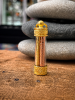 Gold-toned decorative glass capsule with textured gold caps, containing two engraved metal rods, resting on a wooden surface with smooth stones in the background.