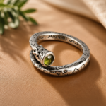 Silver spiral ring with a green faceted gemstone and engraved symbols on orange satin fabric.