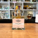 Perfume bottle with a pink label reading AMANDE - MIMOSA, on a wooden counter with blurred store shelves in the background.