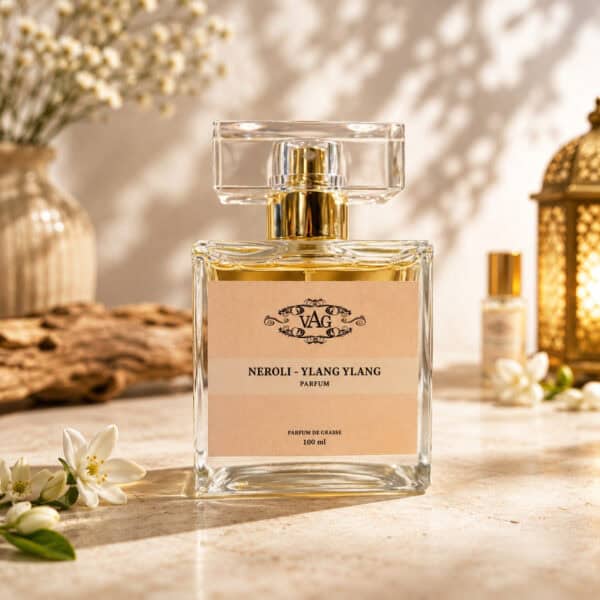 Clear glass perfume bottle with a gold cap and a pastel pink label that reads Neroli - Ylang Ylang, 100 ml, on a sunny countertop with decorative decor in the background.