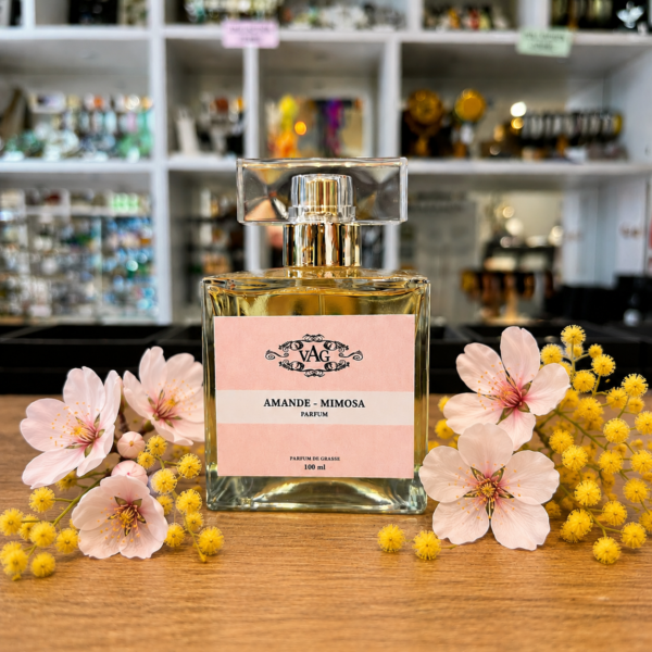 Perfume bottle with a pink label reading Amande - Mimosa, surrounded by pink blossoms and yellow mimosa flowers on a wooden surface