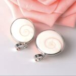 Two round shell pendants with silver frames, featuring white spiral shells, resting on a soft pink surface with rose petals nearby.