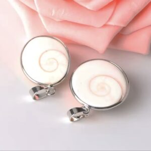 Two round shell pendants with silver frames, featuring white spiral shells, resting on a soft pink surface with rose petals nearby.