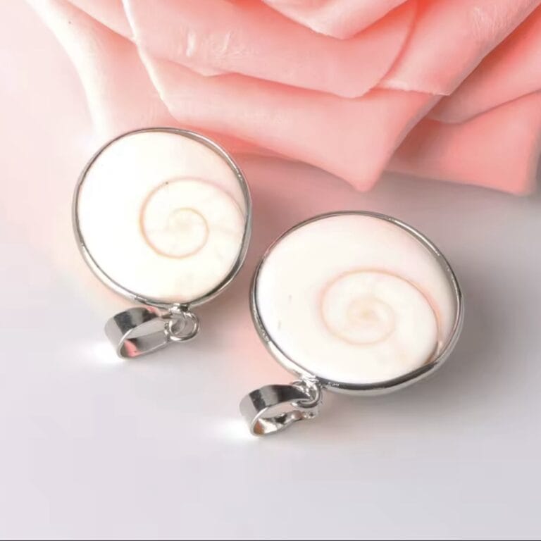 Two round shell pendants with silver frames, featuring white spiral shells, resting on a soft pink surface with rose petals nearby.