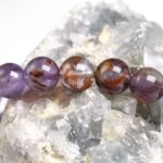 Beaded bracelet of translucent purple amethyst with red-orange inclusions laid on clear quartz crystals