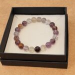 Beaded bracelet with translucent purple, pink, and amber beads arranged in a circle inside a black jewelry box on a white tray.