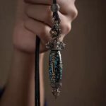 Bracelet/necklace pendant held by hand, featuring an elongated turquoise-inlaid bead with ornate silver filigree details on a black cord.