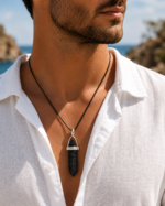 Close-up of a man wearing a black string necklace with a pointed black stone pendant over a white shirt, outdoors by the coast.