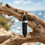 Silver pendant with a black lava stone hanging from weathered driftwood against a blue seascape background