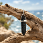 Silver pendant with a black lava stone hanging from weathered driftwood against a blue seascape background